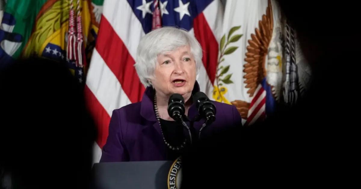 Janet Yellen in a purple shirt speaking at the podium.